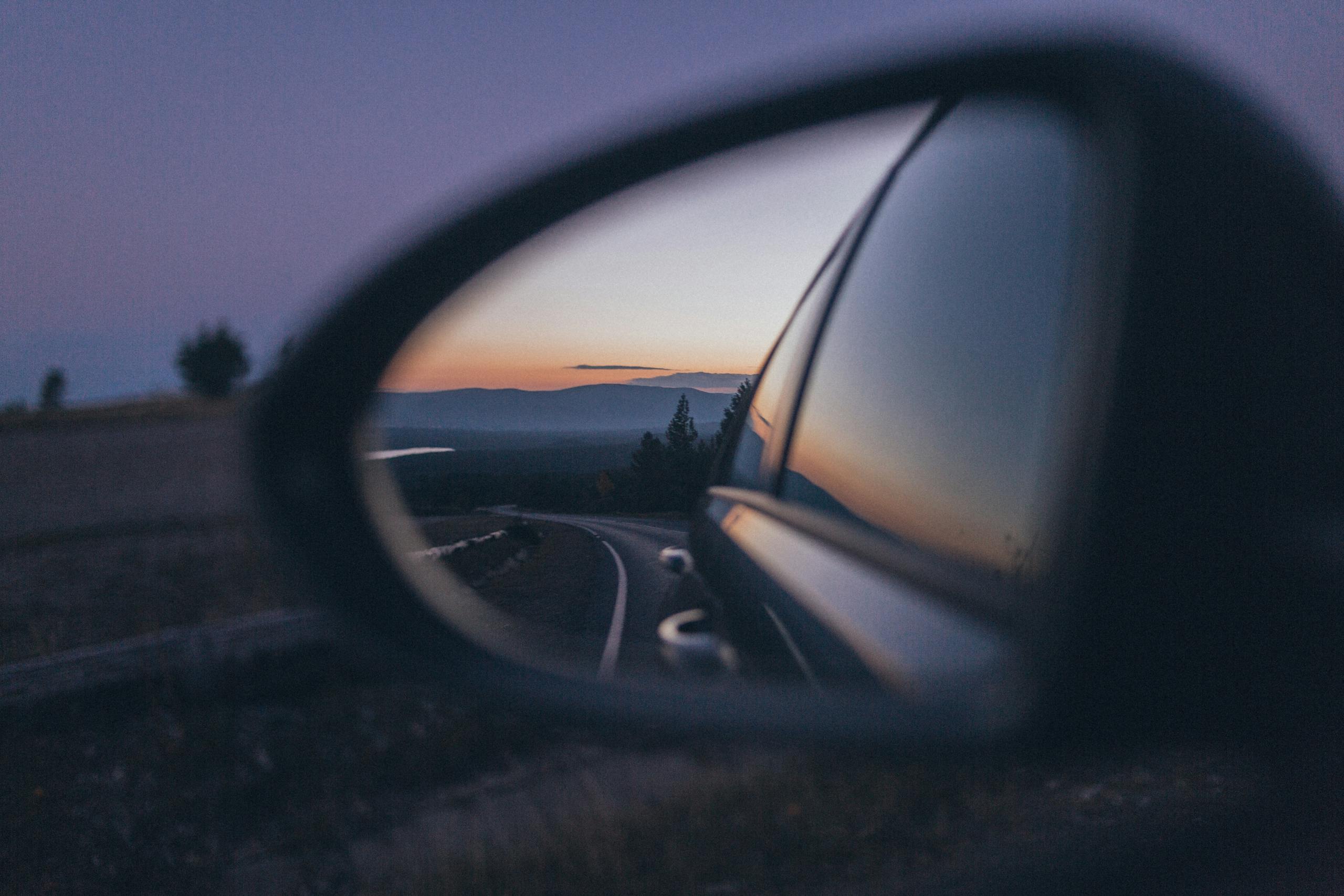 Capture of a scenic sunset reflected in a car's side mirror on a winding road.